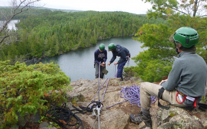 In the foreground, one person wearing safety gear rests on a rock, watching two others on the edge of a cliff. They are also wearing safety gear and are secured by ropes. One appears to be an instructor, giving direction to a student.